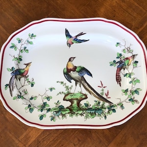 May include: An oval serving platter with a white background and a red scalloped border. The platter features colorful illustrations of birds and foliage. The birds are perched on branches and a mushroom-shaped base.