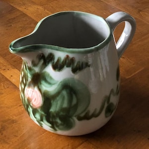 May include: A white ceramic pitcher with a green rim and handle. The pitcher is decorated with green and pink floral designs. The pitcher has a spout for easy pouring and is likely used for serving liquids.