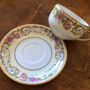 Puede incluir: Un juego de taza y platillo de t&eacute; vintage con un dise&ntilde;o floral. La taza y el platillo son blancos con un borde dorado y presentan un delicado patr&oacute;n floral en tonos de rosa, morado y verde.