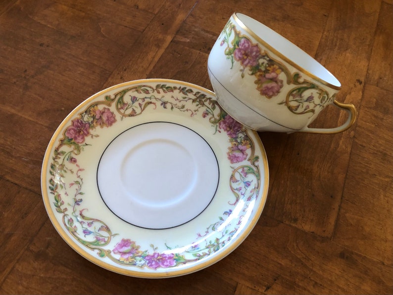 Puede incluir: Un juego de taza y platillo de t&eacute; vintage con un dise&ntilde;o floral. La taza y el platillo son blancos con un borde dorado y presentan un delicado patr&oacute;n floral en tonos de rosa, morado y verde.