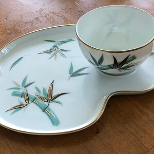 May include: A white porcelain teacup and saucer set with a gold rim and a green and gold bamboo design. The set is on a white porcelain tray with a gold rim and a green and gold bamboo design.