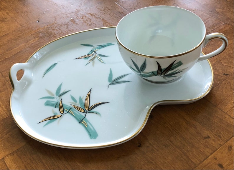 May include: A white porcelain teacup and saucer set with a gold rim and a green and gold bamboo design. The set is on a white porcelain tray with a gold rim and a green and gold bamboo design.