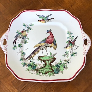 May include: White ceramic serving platter with a scalloped edge and burgundy trim. The platter features a central design of a colorful pheasant and three other birds, surrounded by green foliage.
