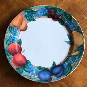 May include: A white ceramic plate with a decorative border featuring a blue and green geometric pattern and illustrations of various fruits, including plums, peaches, and grapes. The plate has a gold rim.