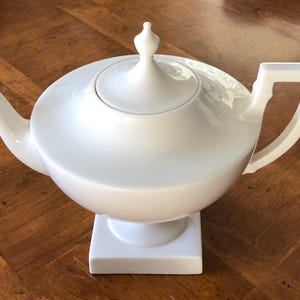May include: A white ceramic teapot with a rounded body, a lid with a small finial, and a square base. The teapot has a curved spout and a rectangular handle. The teapot is displayed on a wooden surface.
