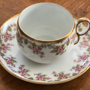 May include: White teacup and saucer set with a floral pattern of pink roses and green leaves. The rims and handle are trimmed with gold. The teacup sits on a matching saucer, creating a classic and elegant look.