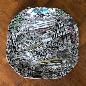 May include: A decorative plate with a black and white transferware design depicting a horse-drawn carriage on a road, passing a church and a half-timbered building. The plate has a scalloped edge and a detailed landscape scene.