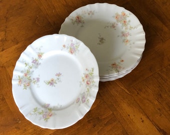 May include: A stack of four white ceramic plates with a delicate floral pattern in shades of pink, purple, and green. The plates have a scalloped edge and are perfect for a formal dinner setting.