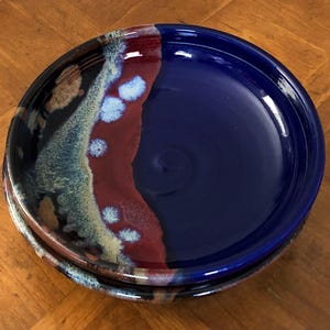 May include: Two ceramic plates stacked on top of each other. The top plate is a deep blue color on the right side and has a mix of red, brown, and blue colors on the left side. The bottom plate is visible.