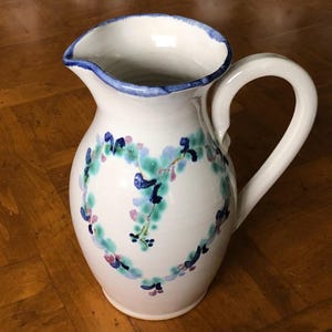 May include: A white ceramic pitcher with a blue rim and a heart-shaped design in shades of blue, green, and pink.