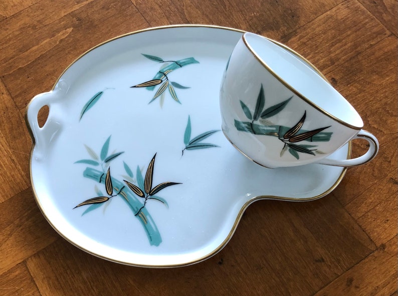 May include: A white porcelain teacup and saucer set with a gold rim and a bamboo design in shades of green and gold.
