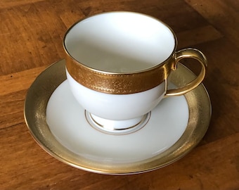 May include: A white teacup and saucer with a gold rim and intricate floral pattern. The cup and saucer are made of fine china.