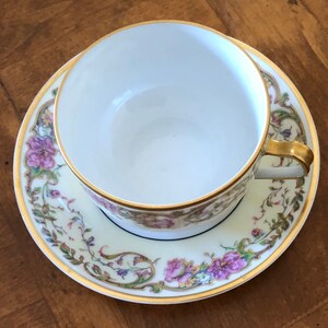 Puede incluir: Una taza de t&eacute; y un platillo de porcelana blanca con un patr&oacute;n floral en tonos de rosa, verde y dorado. La taza y el platillo tienen un borde dorado.