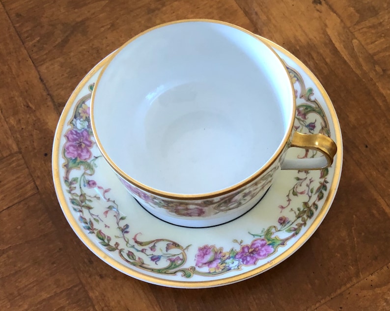 Puede incluir: Una taza de t&eacute; y un platillo de porcelana blanca con un patr&oacute;n floral en tonos de rosa, verde y dorado. La taza y el platillo tienen un borde dorado.