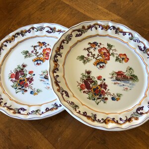 May include: Two white ceramic dinner plates with a scalloped edge and a floral design featuring red, yellow, and blue flowers, green leaves, and a brown vase. The plates have a gold and purple border.