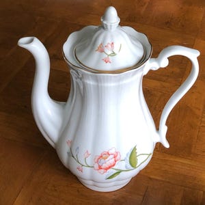 May include: White porcelain coffee pot with a lid, featuring a floral design in pink, orange, and green. The pot has a curved spout and handle, with gold trim around the lid. The pot is approximately 9 inches tall.