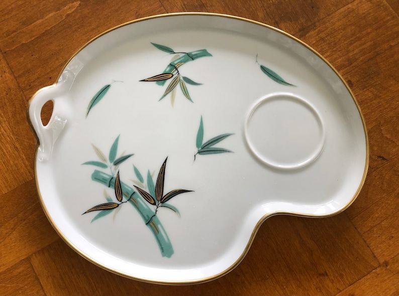 May include: White ceramic serving tray with a gold rim and a green and gold bamboo design. The tray has a small, round indentation for a cup.