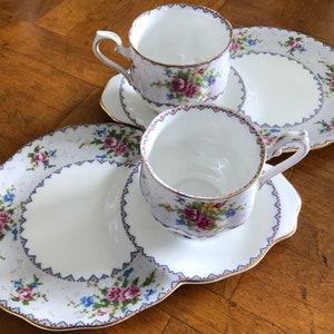 May include: Two white teacups and saucers with floral patterns and gold trim. The saucers are on a larger white plate with a floral pattern and gold trim.