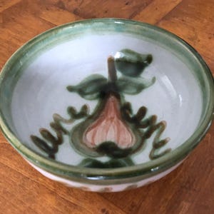 May include: A small, round ceramic bowl with a white interior and a green rim. The bowl features a hand-painted pear design in shades of brown and green. The bowl is likely used for serving small portions of food.