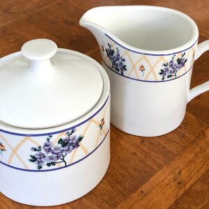 May include: White ceramic sugar bowl and creamer set with a floral design. The design features purple flowers, yellow accents, and a blue and yellow geometric pattern.