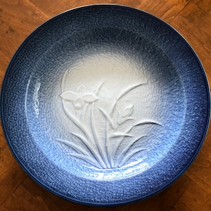 May include: A large, round, blue and white ceramic serving plate with a raised design of a flower and leaves. The plate has a textured, crackled glaze.