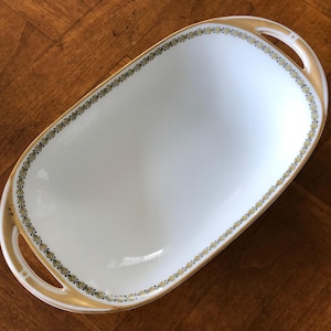 May include: A white oval serving platter with gold trim and handles. The rim is decorated with a delicate pattern of gold and green leaves. The platter is empty and sits on a wooden surface.