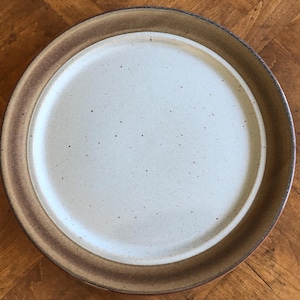 May include: A large, round, white ceramic plate with a brown rim. The plate has a speckled surface and is perfect for serving a variety of dishes.