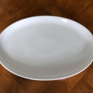 May include: A white, oval-shaped serving platter. The plate has a smooth, glossy surface and a slightly raised rim. The background is a wooden surface with a brown, patterned design. This platter is suitable for serving food.