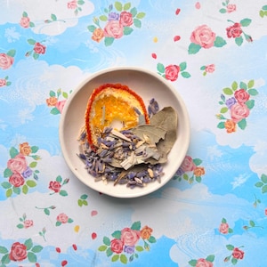 May include: A small, off-white ceramic bowl filled with dried ingredients, including a slice of orange, lavender buds, bay leaves, and other herbs. The bowl sits on a floral patterned fabric with roses and a blue sky background.