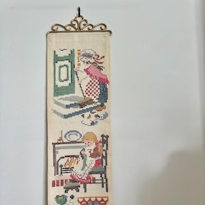 May include: A long, rectangular, vintage cross-stitch wall hanging. The cream-colored fabric features three scenes of people performing domestic tasks. The scenes include a woman by a stove, a girl by a fireplace, and a person using a spinning wheel. A decorative brass hanger is at the top.