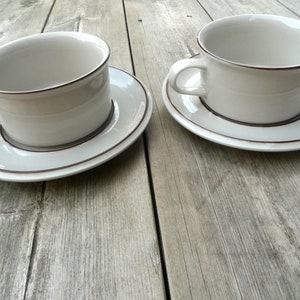 May include: Two white ceramic teacups with brown trim and matching saucers on a wooden surface.