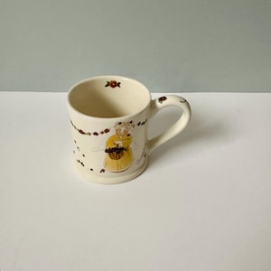 K&ouml;nnte beinhalten: Eine wei&szlig;e Keramiktasse mit einem Blumendesign und einem Comic-Engel, der einen Korb mit Blumen h&auml;lt. Die Tasse hat einen Henkel und ist mit braunen und roten Blumen verziert.