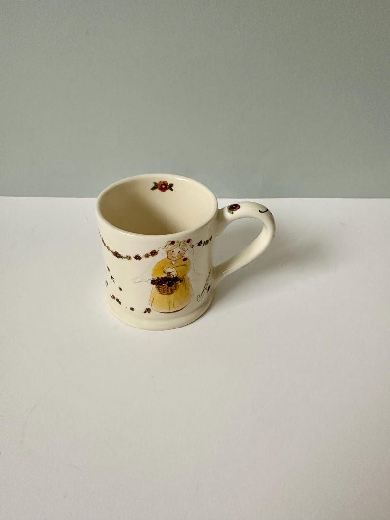 K&ouml;nnte beinhalten: Eine wei&szlig;e Keramiktasse mit einem Blumendesign und einem Comic-Engel, der einen Korb mit Blumen h&auml;lt. Die Tasse hat einen Henkel und ist mit braunen und roten Blumen verziert.