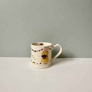 K&ouml;nnte beinhalten: Wei&szlig;e Keramiktasse mit handgemaltem Design einer Frau in einem gelben Kleid, die einen Korb mit Blumen h&auml;lt. Die Tasse hat einen braunen Henkel und ist mit einer Girlande aus Blumen verziert.