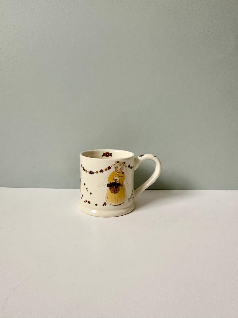 K&ouml;nnte beinhalten: Wei&szlig;e Keramiktasse mit handgemaltem Design einer Frau in einem gelben Kleid, die einen Korb mit Blumen h&auml;lt. Die Tasse hat einen braunen Henkel und ist mit einer Girlande aus Blumen verziert.