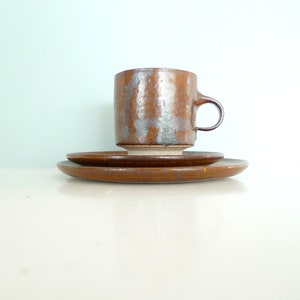 Può includere: Un set da caffè in ceramica marrone composto da una tazza e un piattino. La tazza ha una smaltatura marrone screziata con un tocco di argento. Il piattino è di un marrone uniforme.