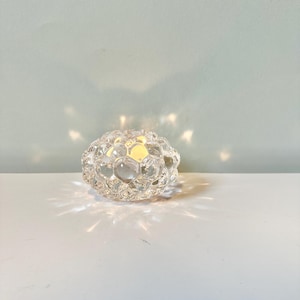 May include: A clear glass votive candle holder with a textured, honeycomb-like surface. The glass features multifaceted, rounded indentations that catch and refract light, creating a glowing effect with distinct light rays radiating outward.