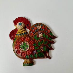 Könnte beinhalten: Ein Keramik-Hahn zum Aufhängen an der Wand mit braunem Untergrund und bunten Blumen- und Blattmotiven. Der Hahn hat einen roten Kamm, einen gelben Schnabel und blaue Augen.