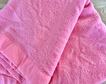 Op de afbeelding: Een roze fleece deken met een satijnen rand. De deken is dubbelgevouwen en ligt op een houten oppervlak.