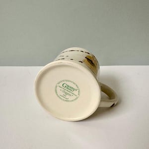 K&ouml;nnte beinhalten: Wei&szlig;e Keramiktasse mit braunem und goldenem Design. Am Boden der Tasse befindet sich ein gr&uuml;nes Etikett mit der Aufschrift "Gnomes' Garden" und "Angels of the Month Collection by Countryside" mit einem Copyright-Symbol.