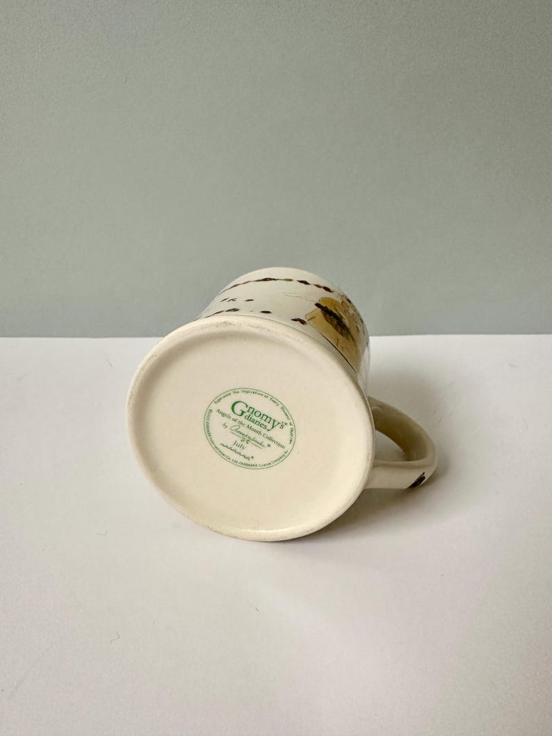 K&ouml;nnte beinhalten: Wei&szlig;e Keramiktasse mit braunem und goldenem Design. Am Boden der Tasse befindet sich ein gr&uuml;nes Etikett mit der Aufschrift "Gnomes' Garden" und "Angels of the Month Collection by Countryside" mit einem Copyright-Symbol.