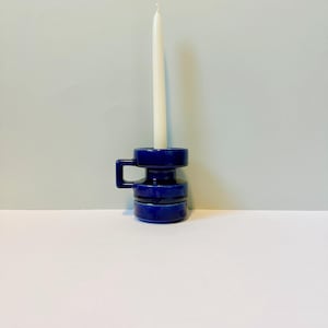 Könnte beinhalten: Ein dunkelblauer Keramik-Kerzenhalter mit Griff, der eine hohe, weiße Kerze hält. Der Kerzenhalter hat ein zylindrisches Design mit horizontalen Rillen. Der Hintergrund ist hellblau und weiß.