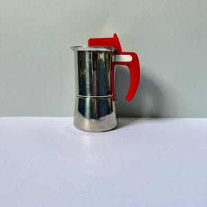 Può includere: Una caffettiera in acciaio inossidabile con manico e parte superiore rosso brillante. La caffettiera ha un design classico ed è realizzata in metallo lucido. Il manico è curvo per una facile presa. Progettata per preparare l'espresso sul fornello.