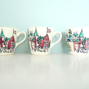 May include: Three white ceramic mugs with a colorful illustration of a town and the word "VARDE" printed on the front.