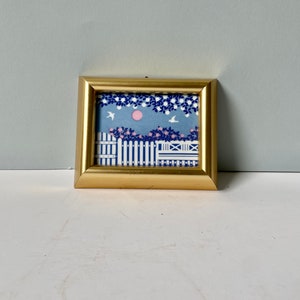 May include: A small framed print with a gold-colored frame. The artwork depicts a blue sky with a pink sun, white birds, and a white picket fence with pink flowers. The frame is square.