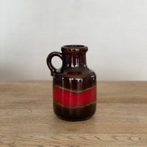Peut inclure: Un vase en céramique brun avec un glaçage rouge et brun. Le vase a une anse et un col étroit.