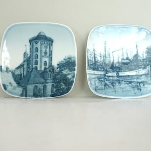 May include: Two square ceramic plates with blue and white designs. One plate features a building with a tower, while the other depicts a harbor scene with boats.
