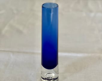 Op de afbeelding: Een lange, cilindrische glazen vaas met een verloop van diepblauw naar helder. De vaas heeft een afgeronde basis en een smalle opening, geschikt voor een enkele bloem of een klein boeket. De vaas is ongeveer 20 cm hoog.