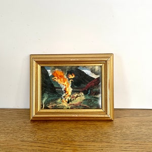 May include: A rectangular painting in a gold-toned frame depicting a landscape scene with a large, bright orange and yellow fire burning in the center. Dark mountains rise in the background, and a group of small figures stands near the flames.