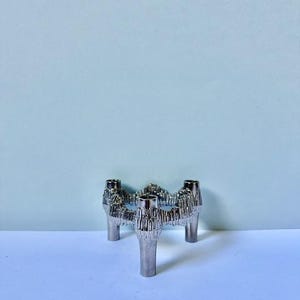 May include: A silver-colored, three-legged candle holder with four candle sockets. The candle holder has a textured, organic design and is set against a light blue background.
